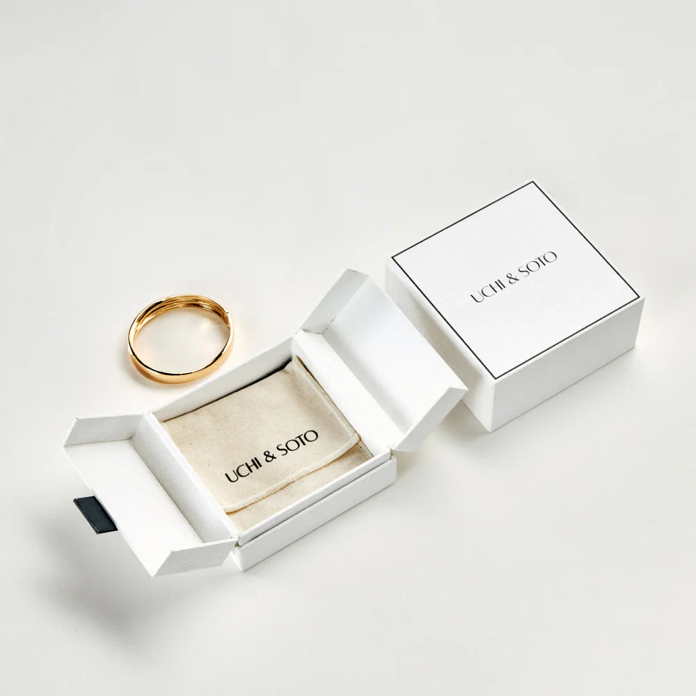 Gold bangle in an open white box with 'UCHI & SOTO' branding on a light gray background
